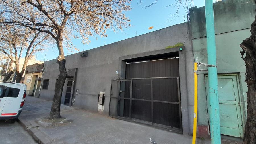 Frente de los galpones de Beazley 3549: portón de chapa oscuro sobre un muro gris, con un árbol en la vereda