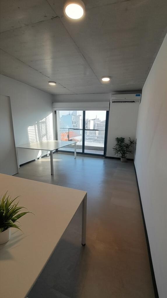 Oficina despejada y ambientada, vista desde la entrada: mesas blancas, plantas y, al fondo, un ventanal con salida al balcón (imagen modificada con IA)